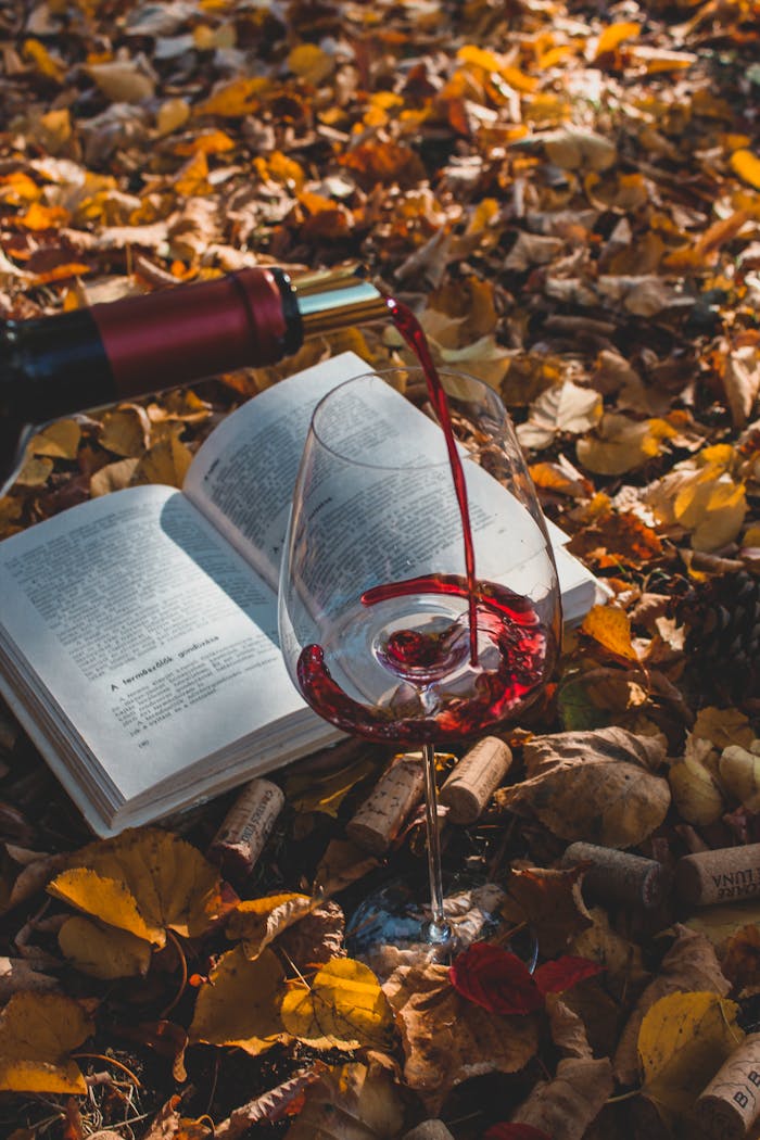 Red wine pouring into glass amidst autumn leaves, open book and corks.